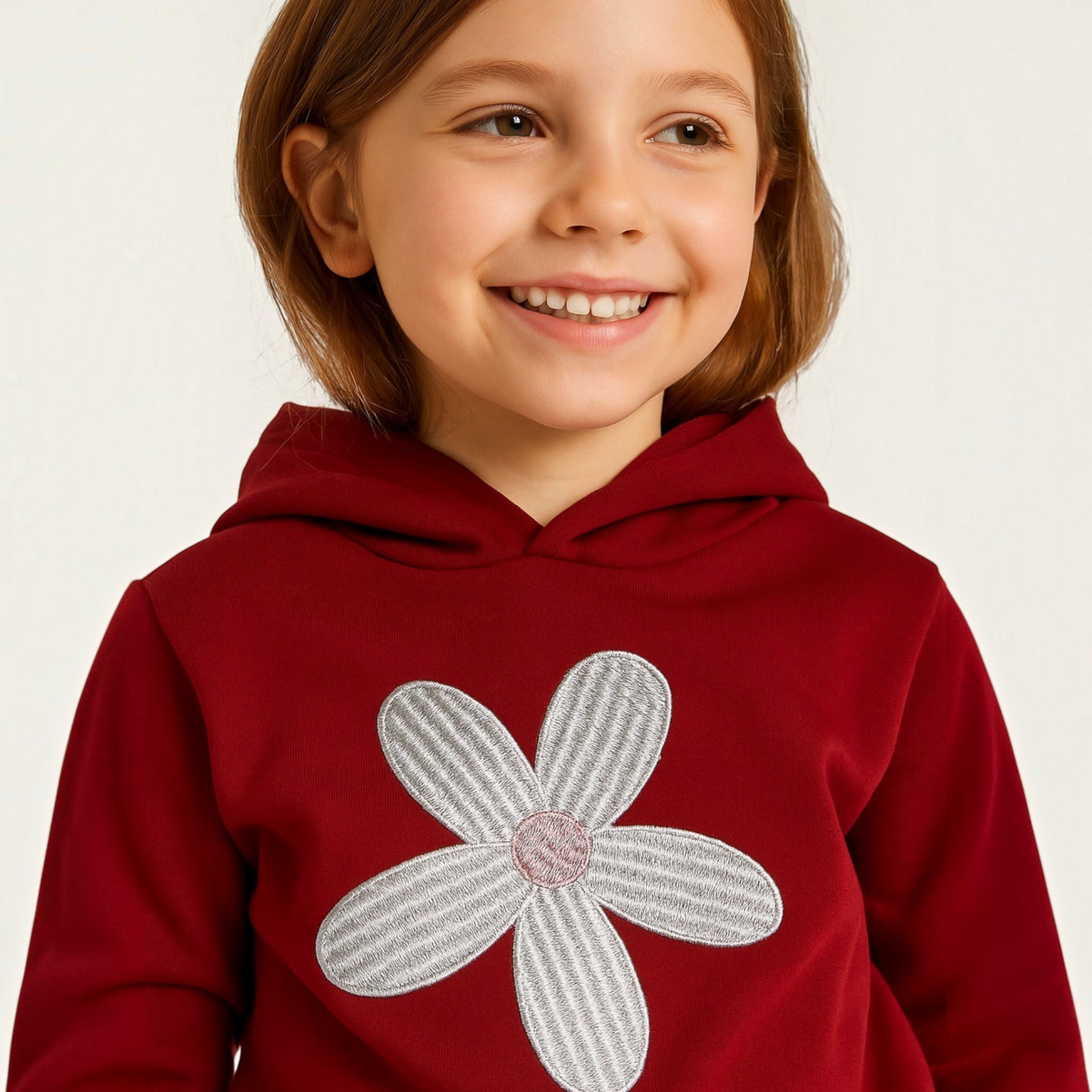 Young girl smiling while wearing a deep red victor and jane hoodie featuring a large white daisy flower applique.
