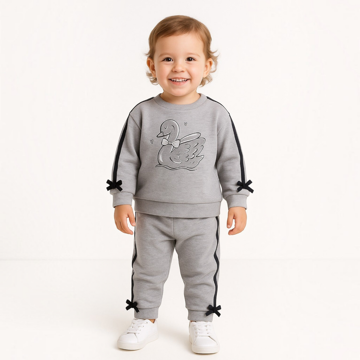 A toddler wearing a grey victor and jane 2-piece embroidered set with a swan graphic and black bow accents.