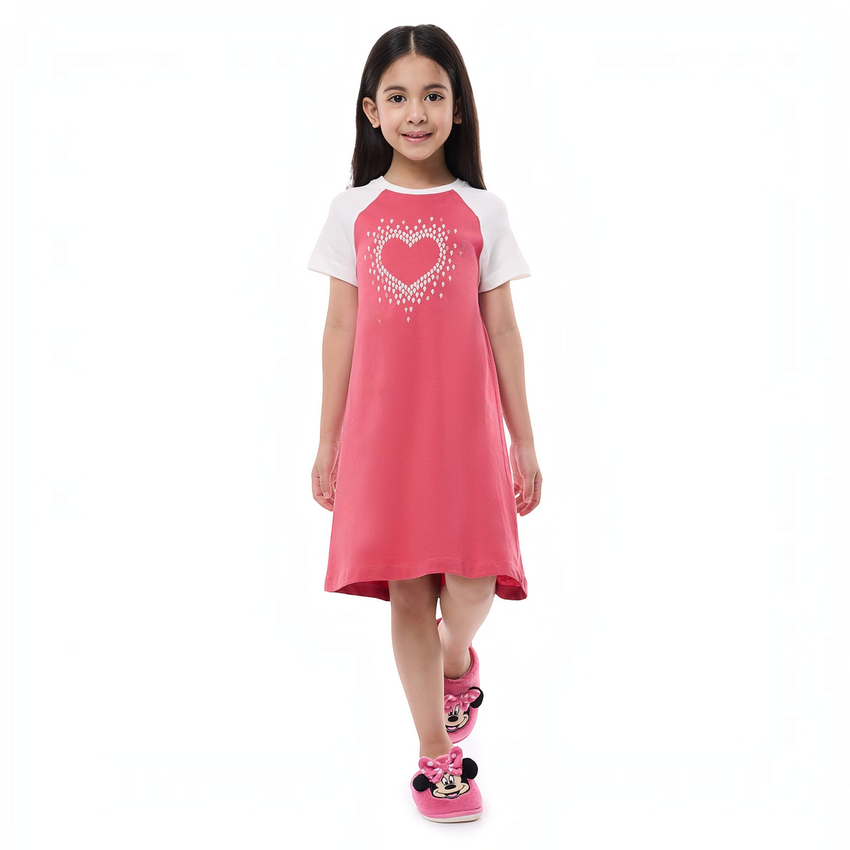 A young girl wearing a victor and jane pink and white short-sleeve pajama dress with a heart graphic and slippers.