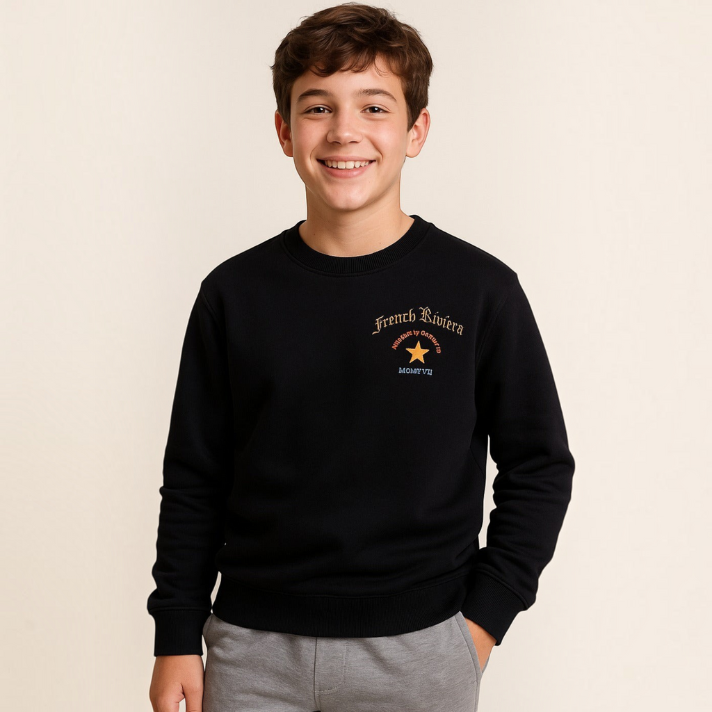 A smiling boy wearing a black victor and jane graphic sweatshirt with French Riviera text and a star design.