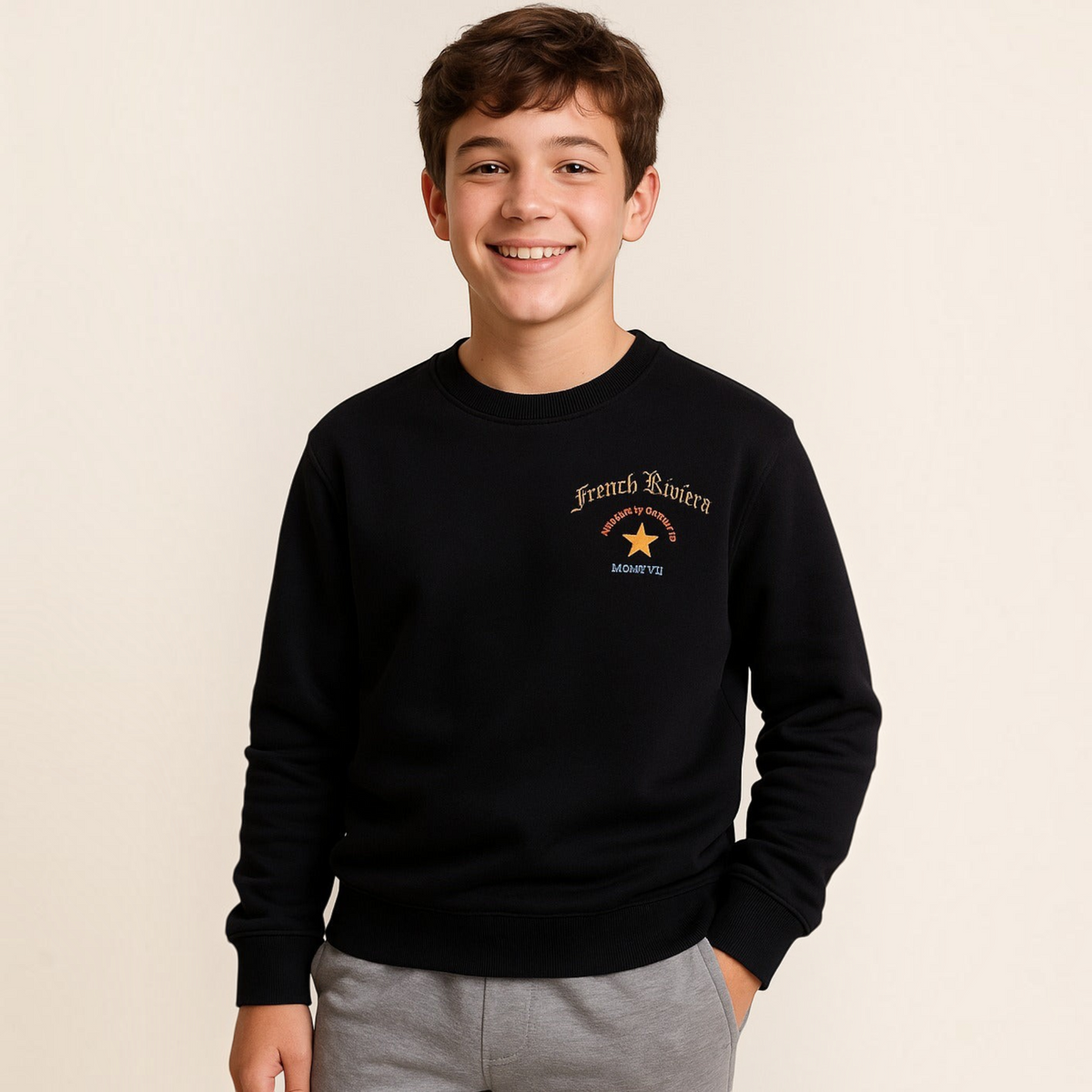 A smiling boy wearing a black victor and jane graphic sweatshirt with French Riviera text and a star design.