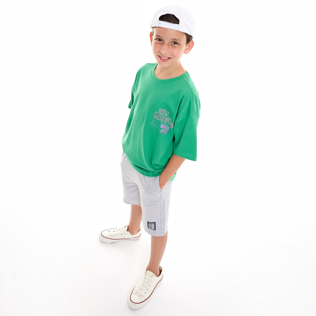 A young boy wearing a green victor and jane graphic t-shirt, grey shorts, white cap, and white sneakers.