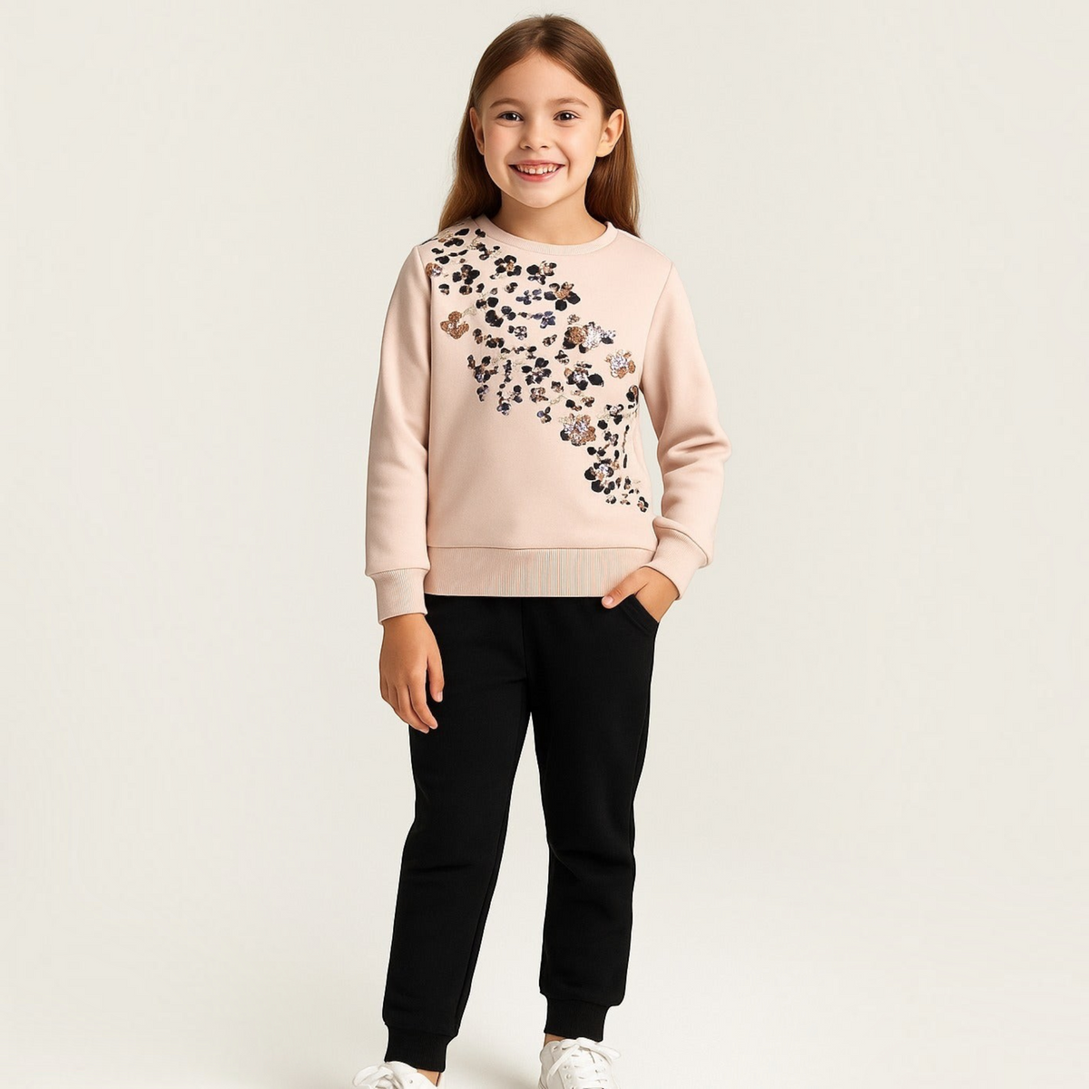Young girl wearing a victor and jane light pink sweatshirt with floral sequin detailing and black joggers