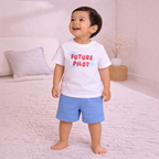 A toddler boy wearing a victor and jane white cotton t-shirt with Future Pilot text and blue shorts in a bedroom.