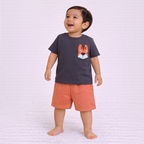 A toddler boy wearing a victor and jane charcoal grey t-shirt with a tiger pocket and orange shorts.