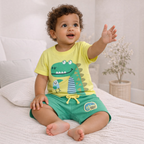 A baby boy wearing a victor and jane yellow dinosaur t-shirt and green shorts with a jungle vibes patch