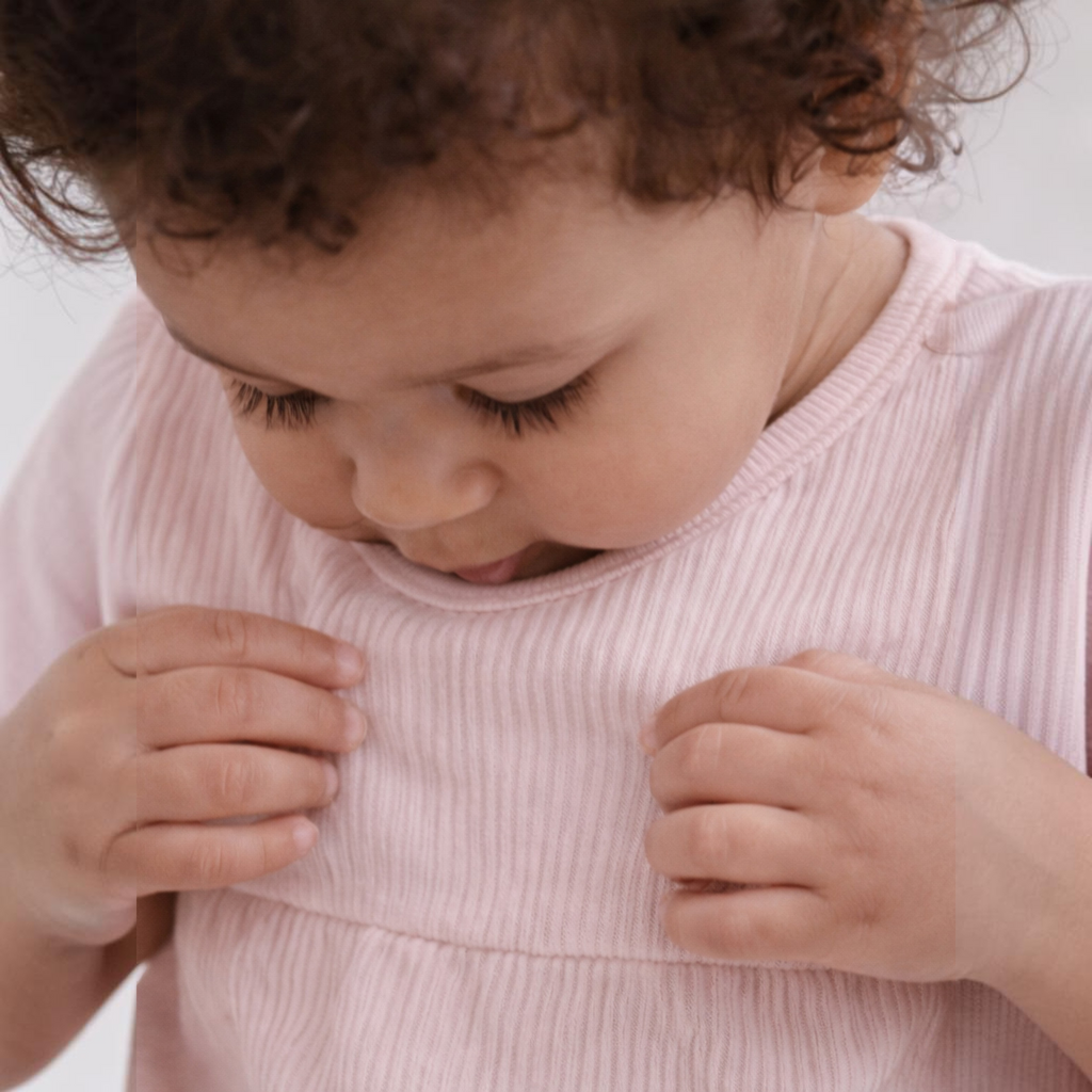 A toddler wearing a soft pink ribbed shirt from victor and jane, looking down with hands touching the fabric.