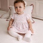 Baby girl wearing a victor and jane pink floral print dress with matching white ribbed tights while sitting on a rug.
