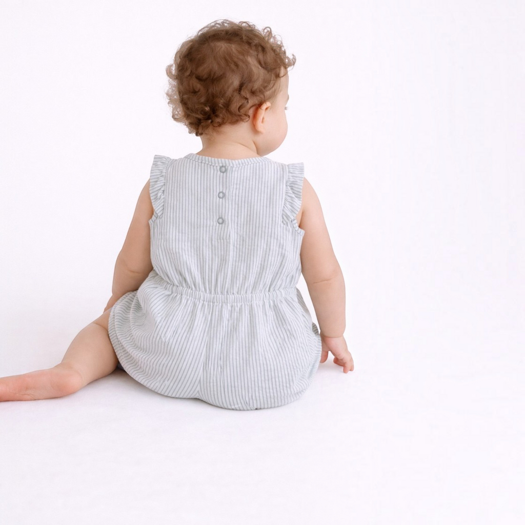 Baby girl wearing a victor and jane light grey striped sleeveless romper with ruffled shoulders, back view