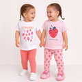 Two girls wearing victor and jane strawberry graphic t-shirt and leggings sets in white, pink, and coral colours.