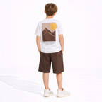 A boy wearing a victor and jane white graphic t-shirt with a mountain print and brown shorts from behind.