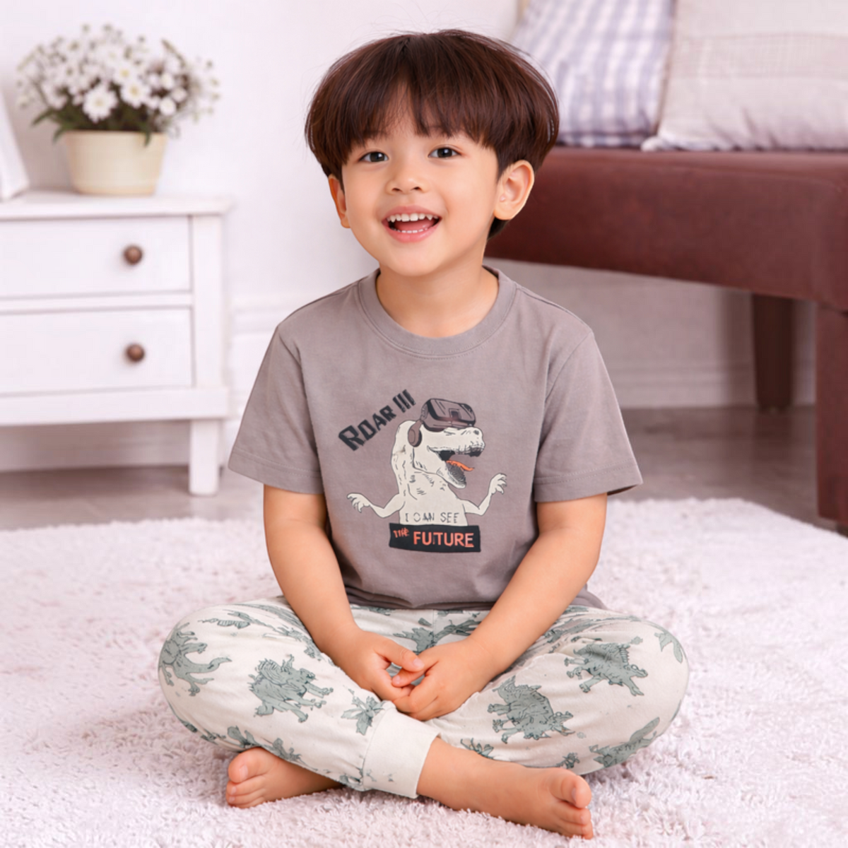A young boy wearing a victor and jane grey graphic t-shirt with a dinosaur print and matching patterned pyjama pants.