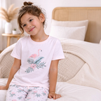 Young girl wearing a victor and jane light pink flamingo print t-shirt and floral pajama bottoms on a bed