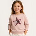 A smiling young girl wearing a light pink victor and jane sweatshirt featuring a large sequined star design.