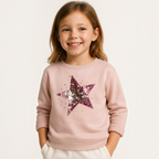 A smiling young girl wearing a light pink victor and jane sweatshirt featuring a large sequined star design.